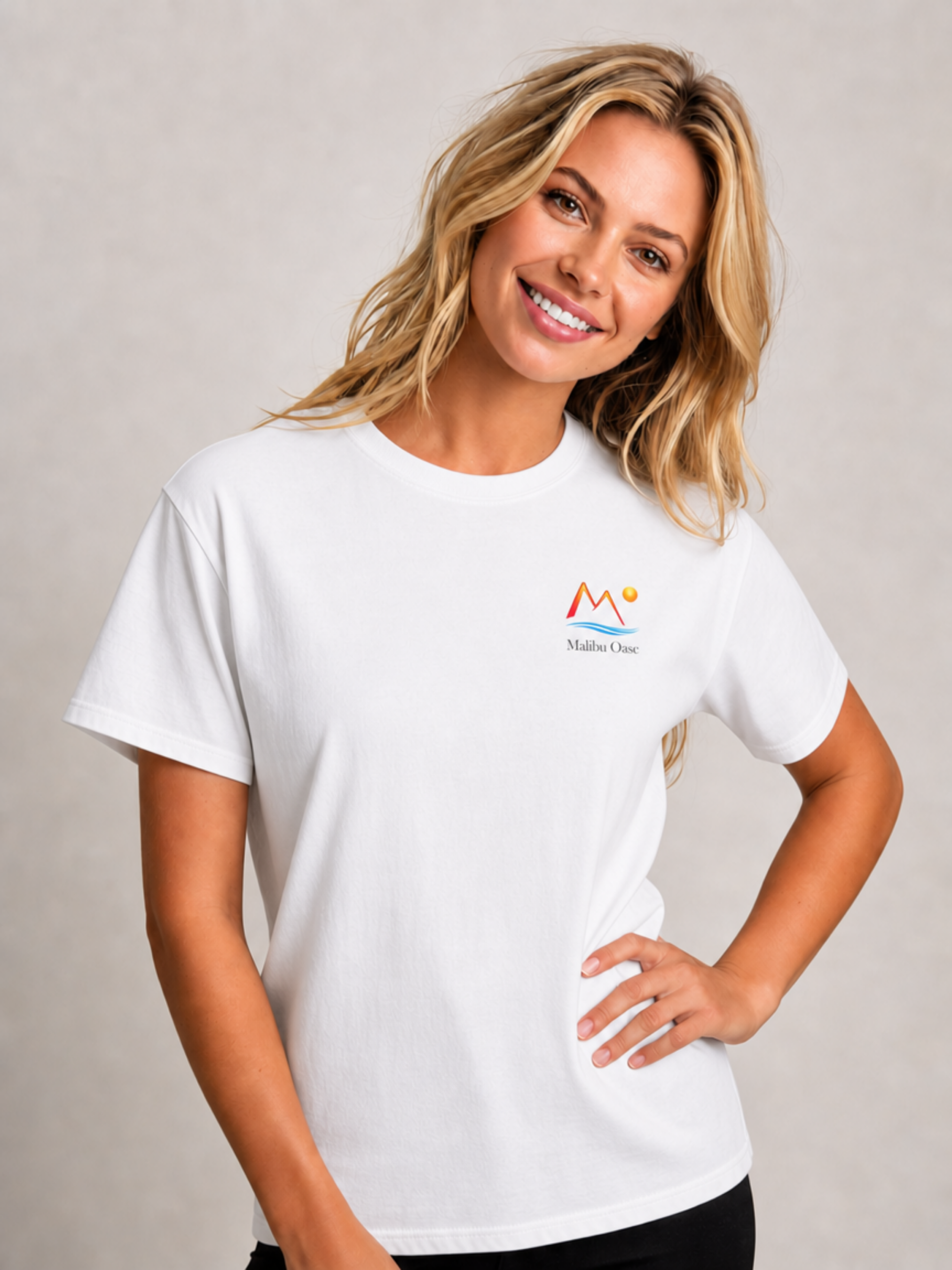 A smiling blonde woman wearing a white short-sleeve crewneck t-shirt with a colorful mountain logo and the text 'Malibu Oasi' on the left chest, posed against a neutral light gray studio background.