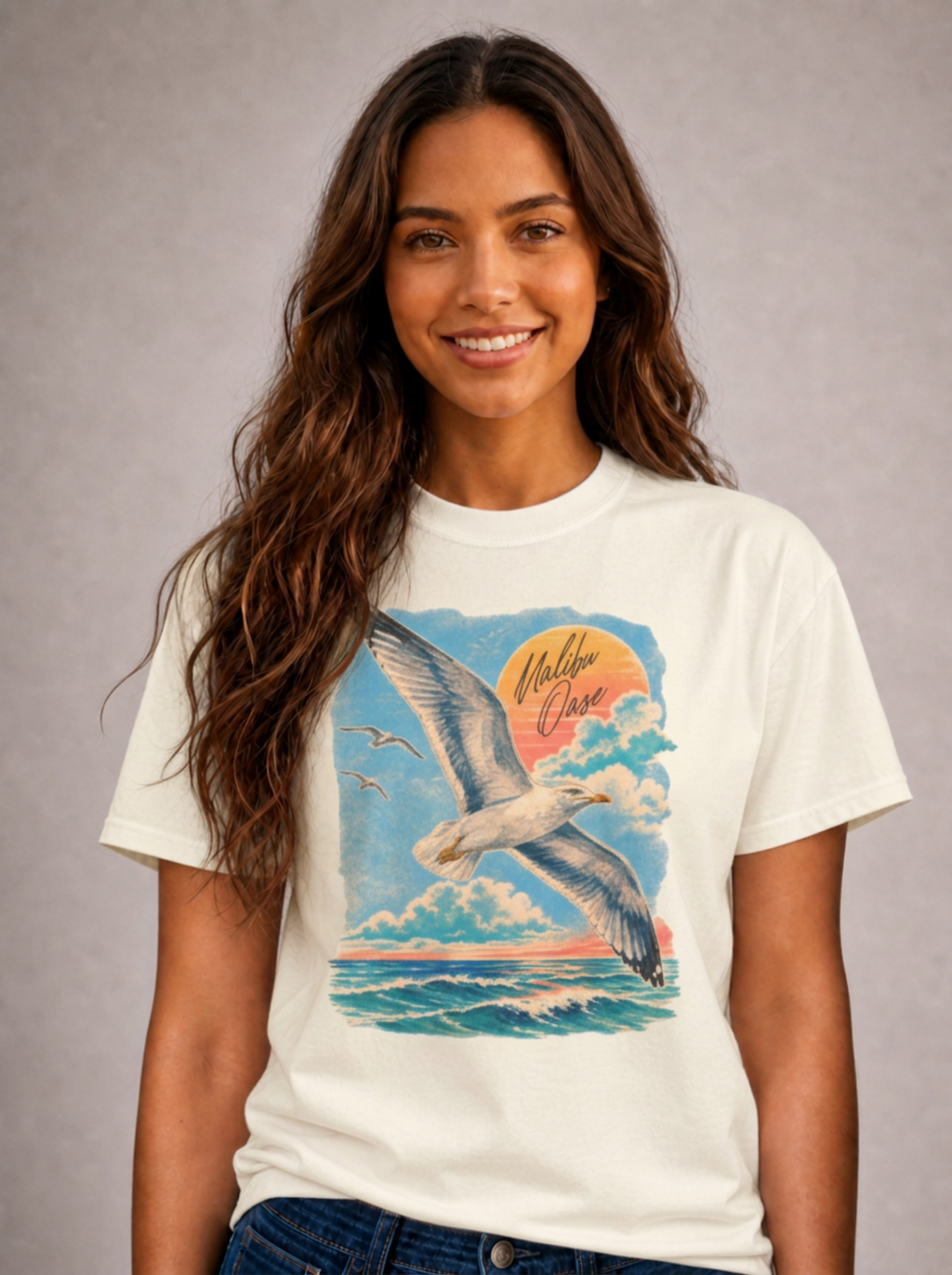 A smiling woman with long wavy brown hair models a cream-colored graphic t-shirt. The t-shirt features a colorful print of a seagull flying over the ocean under a sunset, with the words 'Malibu Oase' written in a cursive font.