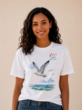 A smiling woman with long curly dark hair wears a white graphic T-shirt featuring a seagull over water and the text/logo 'Malibu Oase.' The photo is taken against a plain beige wall with the model's torso and upper legs visible.
