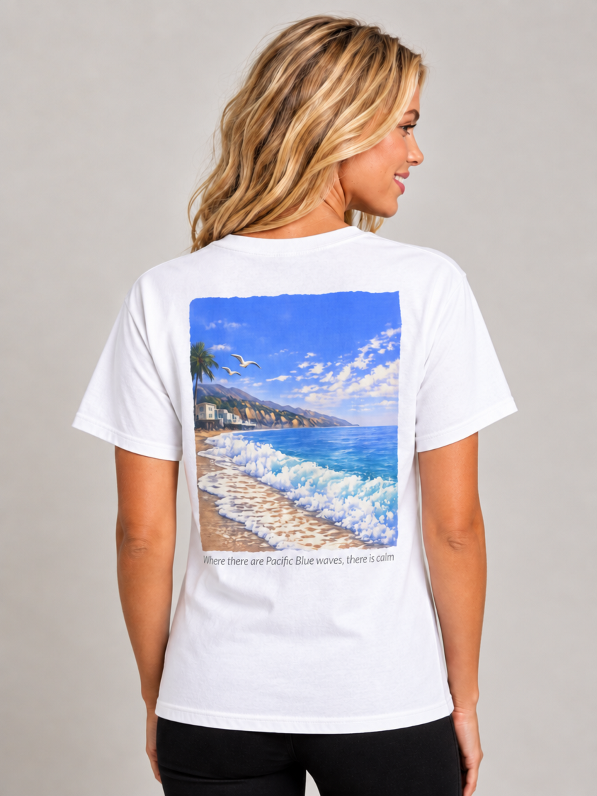 A woman shown from the back wearing a white short-sleeve T-shirt with a large square graphic print of a beach scene and the text 'Where there are Pacific Blue waves, there is calm.'