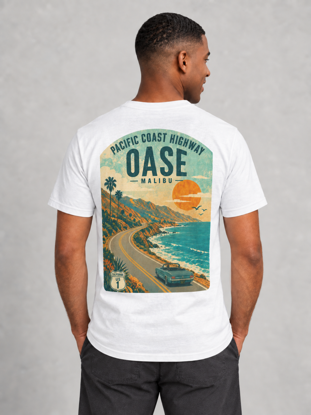 A man wearing a white graphic t-shirt, viewed from the back. The shirt features a large retro-style illustration of the Pacific Coast Highway in Malibu, California, with a vintage car, ocean, and palm trees.
