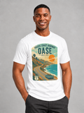 A man wearing a white graphic t-shirt with a vintage-style coastal highway print that reads 'PACIFIC COAST HIGHWAY OASE MALIBU'. The t-shirt features a retro illustration of a winding road, ocean, hills, and a car under a sunset sky.