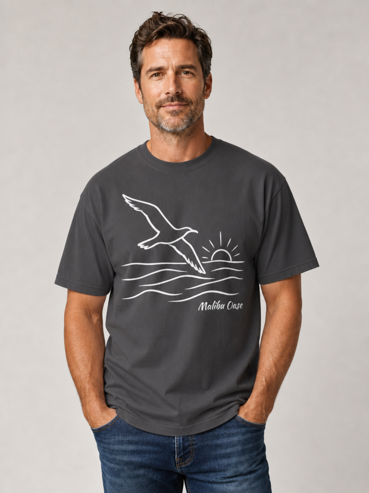 A man wearing a dark gray t-shirt with a white line art graphic of a seagull flying over waves towards a rising sun, with the text 'Malibu Oase' printed below. He is wearing blue jeans and standing against a neutral light gray background.