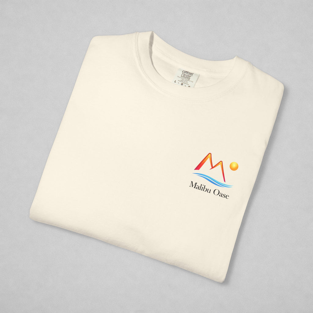 A folded off-white crewneck t-shirt with a colorful 'Malibu Oase' graphic on the left chest, displayed on a neutral gray background.