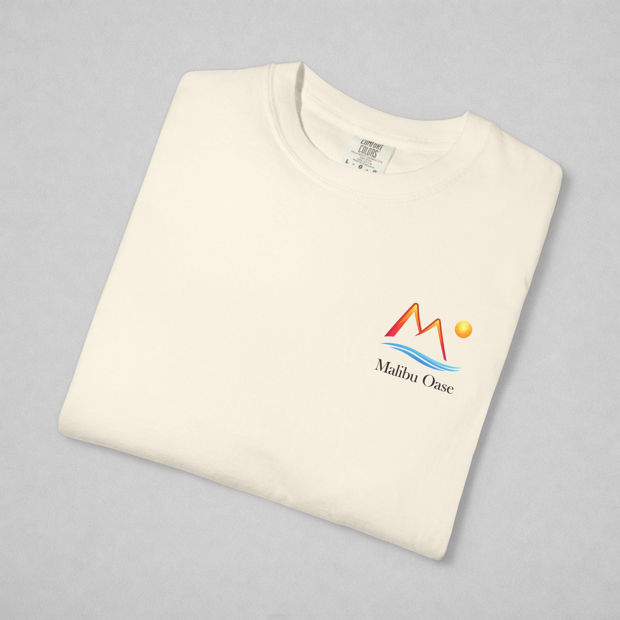 A folded off-white crewneck t-shirt with a colorful 'Malibu Oase' graphic on the left chest, displayed on a neutral gray background.