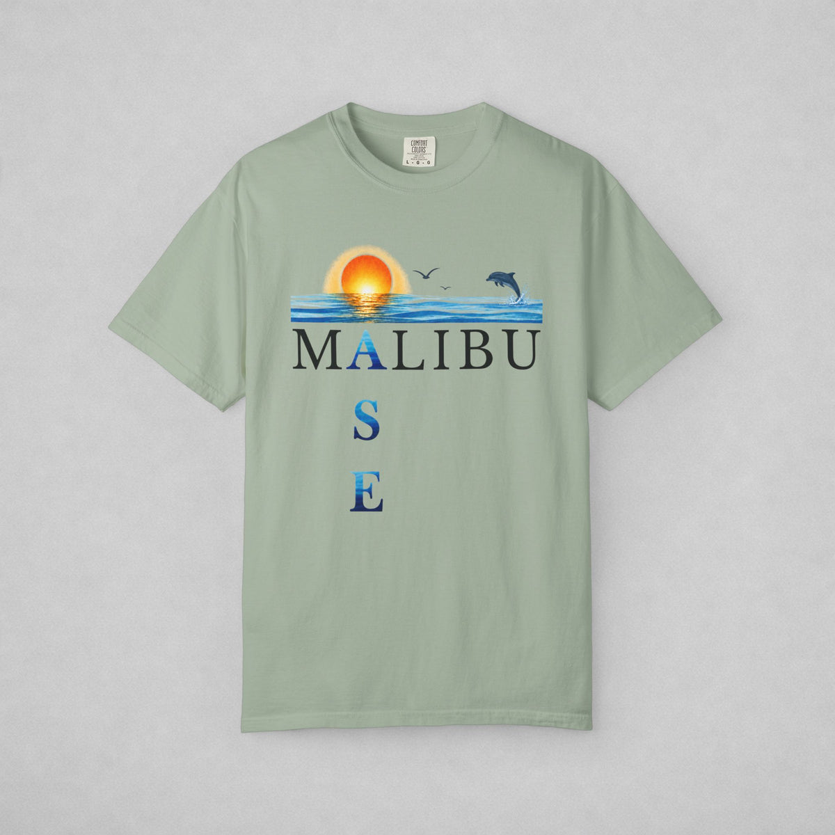 A sage green short-sleeve t-shirt with a 'MALIBU OASE' graphic on the chest featuring a sunset, ocean waves, birds, and a dolphin, displayed flat against a neutral gray background.