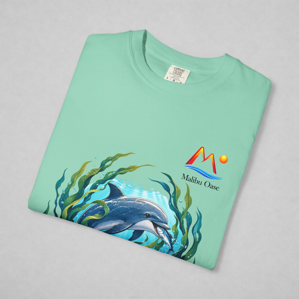 A folded mint green t-shirt on a light gray background, featuring a colorful graphic of a dolphin swimming among green seaweed, with the 'Malibu Oase' logo printed on the chest.