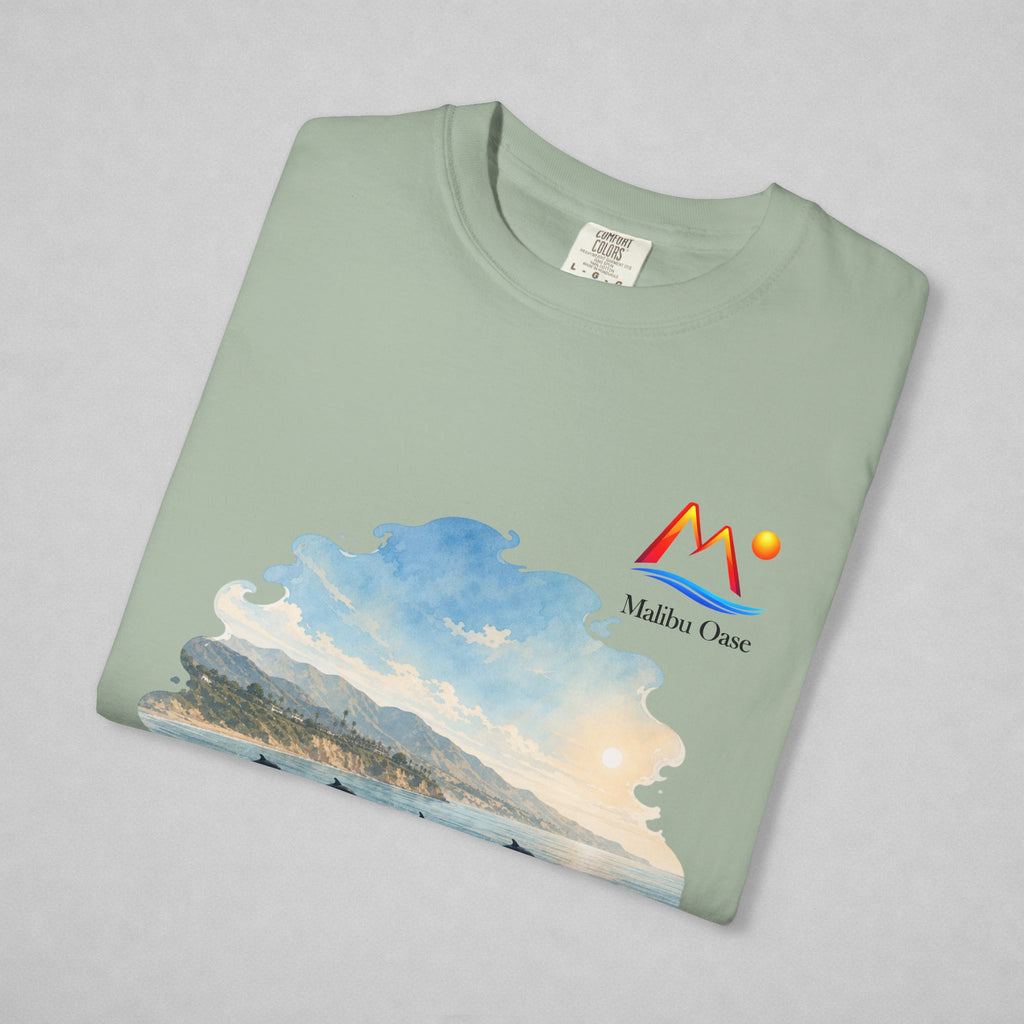 A folded Bay t-shirt on a light gray background, featuring a colorful graphic print of a coastal landscape with mountains, waves, and a sun, alongside the 'Malibu Oase' logo.