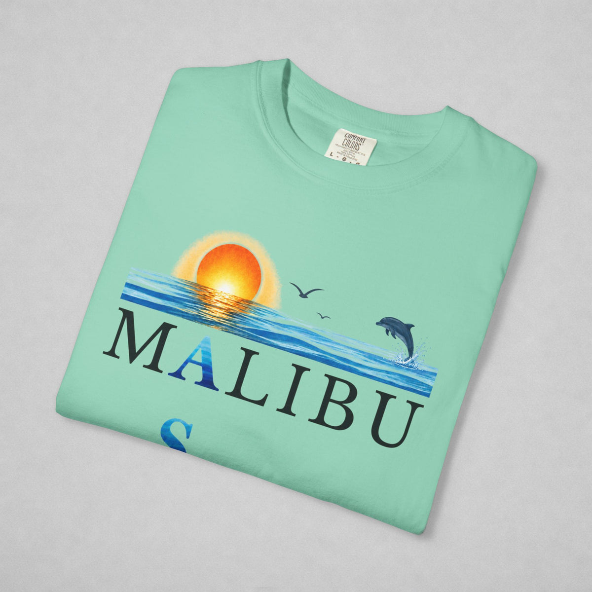 A mint green folded t-shirt with a 'MALIBU' graphic print featuring a sunset, ocean waves, a dolphin, and birds, displayed on a neutral gray background.