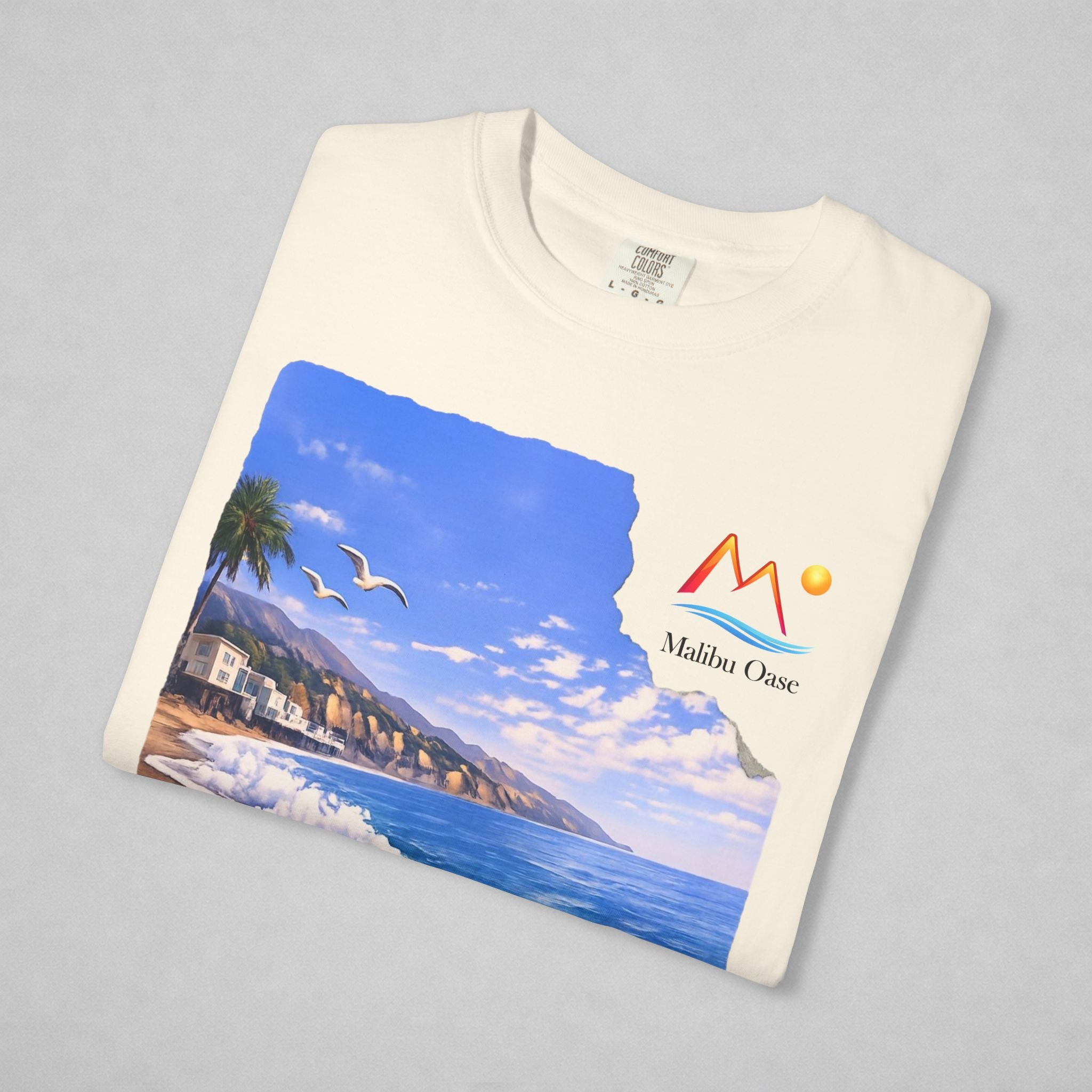 A folded off white t-shirt on a neutral gray background, featuring a printed graphic of a coastal landscape with a mountain logo that says 'Malibu Oase.'