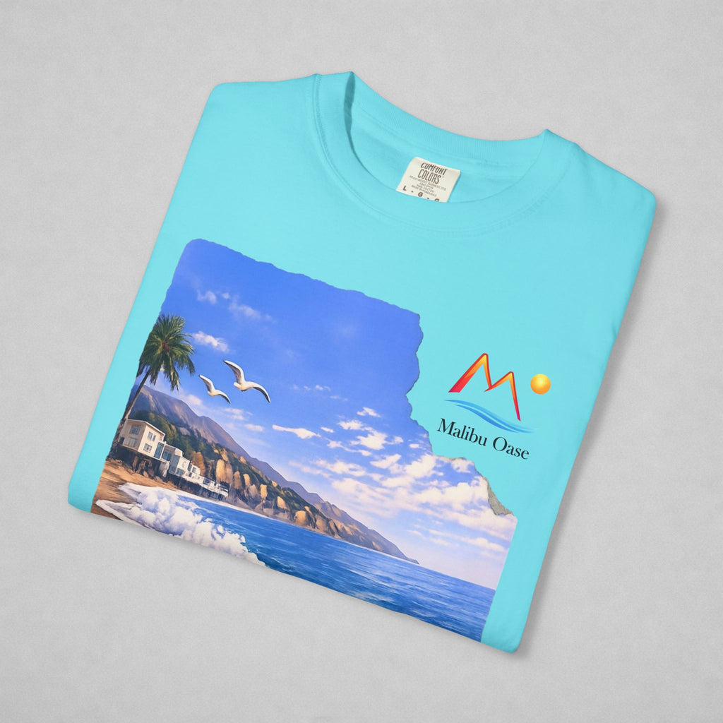 A folded light blue t-shirt on a neutral gray background, featuring a printed graphic of a coastal landscape with a mountain logo that says 'Malibu Oase.'