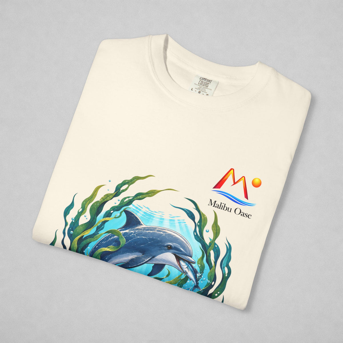 An off-white folded t-shirt on a light gray background, featuring a colorful graphic of a dolphin swimming underwater with green seaweed and the text 'Malibu Oase' next to a stylized red and orange mountain logo.