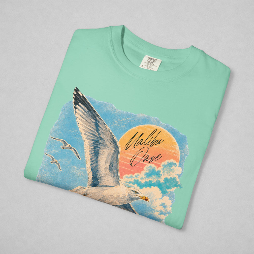 A mint green crewneck t-shirt with a graphic print of a seagull flying past a sunset over clouds, featuring the text 'Malibu Oase'. The shirt is laid flat on a light gray background.