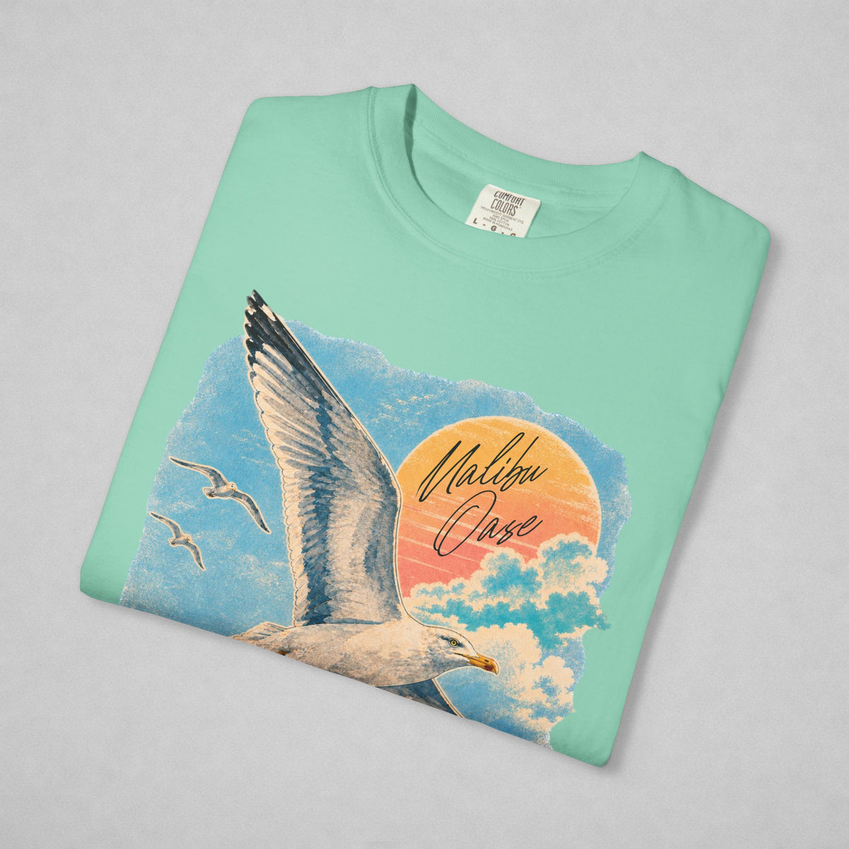 A mint green crewneck t-shirt with a graphic print of a seagull flying past a sunset over clouds, featuring the text 'Malibu Oase'. The shirt is laid flat on a light gray background.