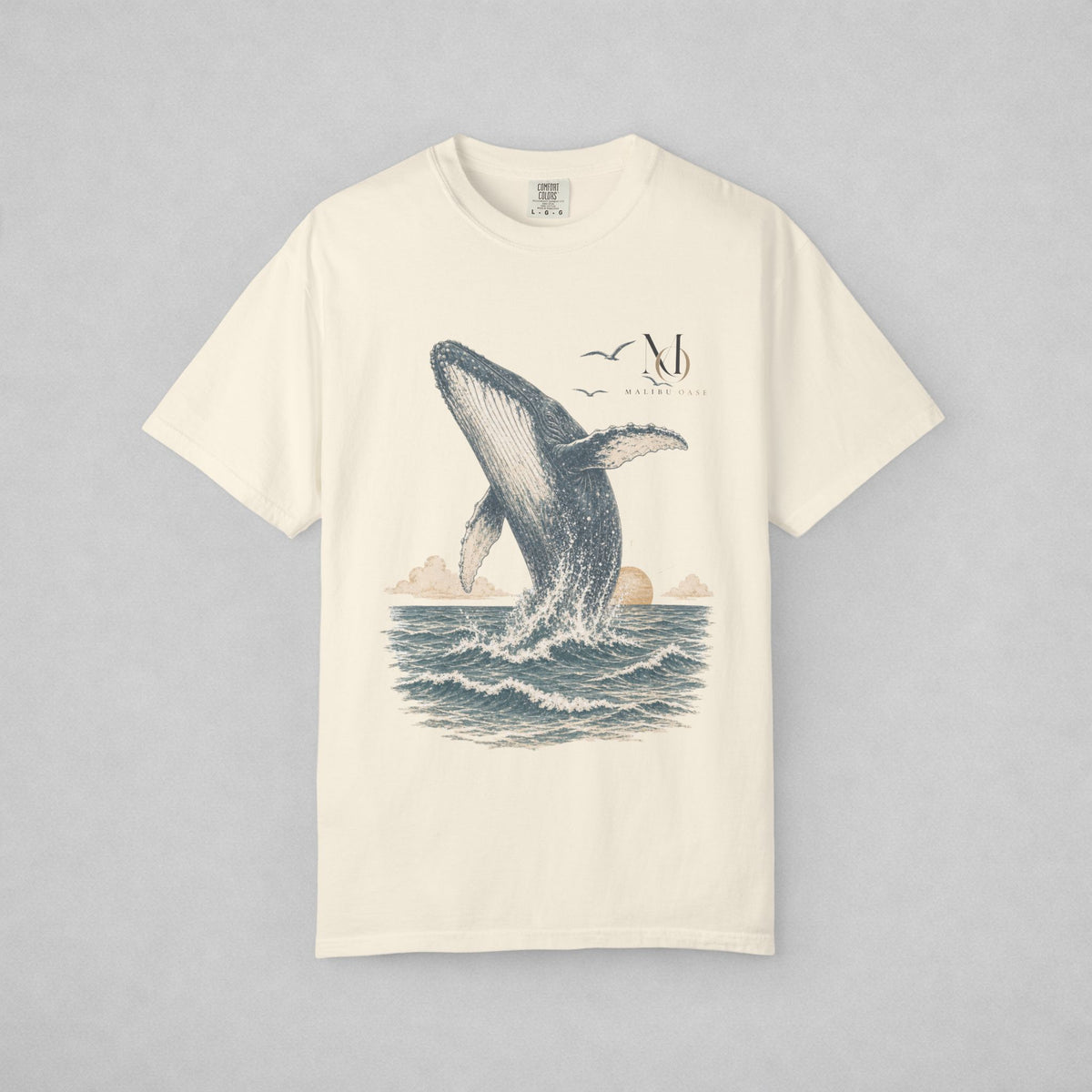An off-white short-sleeve crewneck t-shirt featuring a detailed graphic print of a beluga whale breaching the ocean, with a small stylized 'Malibu Oase' monogram and seagulls in the sky above.