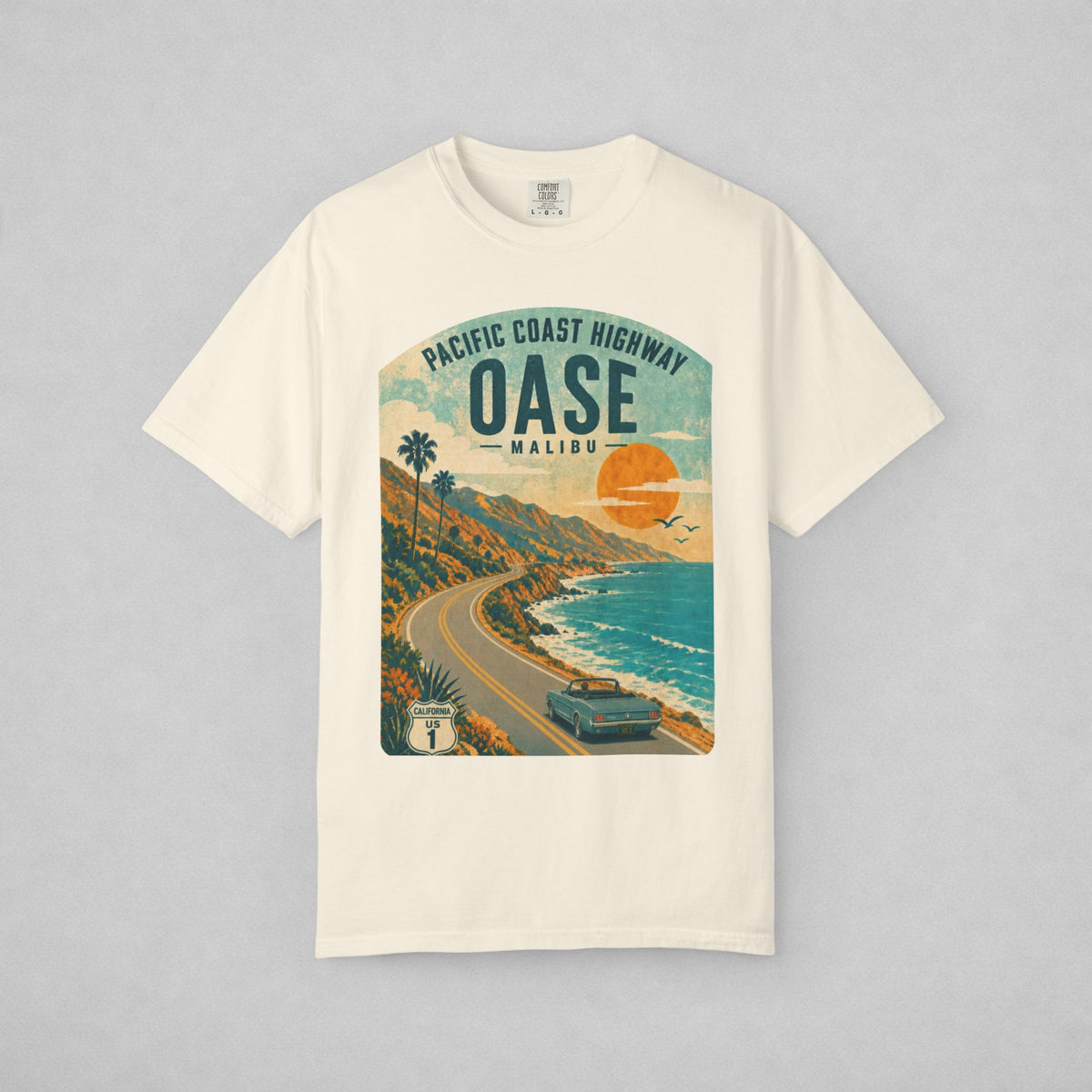 An off-white short-sleeve t-shirt with a vintage-style graphic print of the Pacific Coast Highway in Malibu, California. The graphic features a retro sunset, palm trees, a winding coastal road, and a car, with 'OASE MALIBU' printed in bold text.