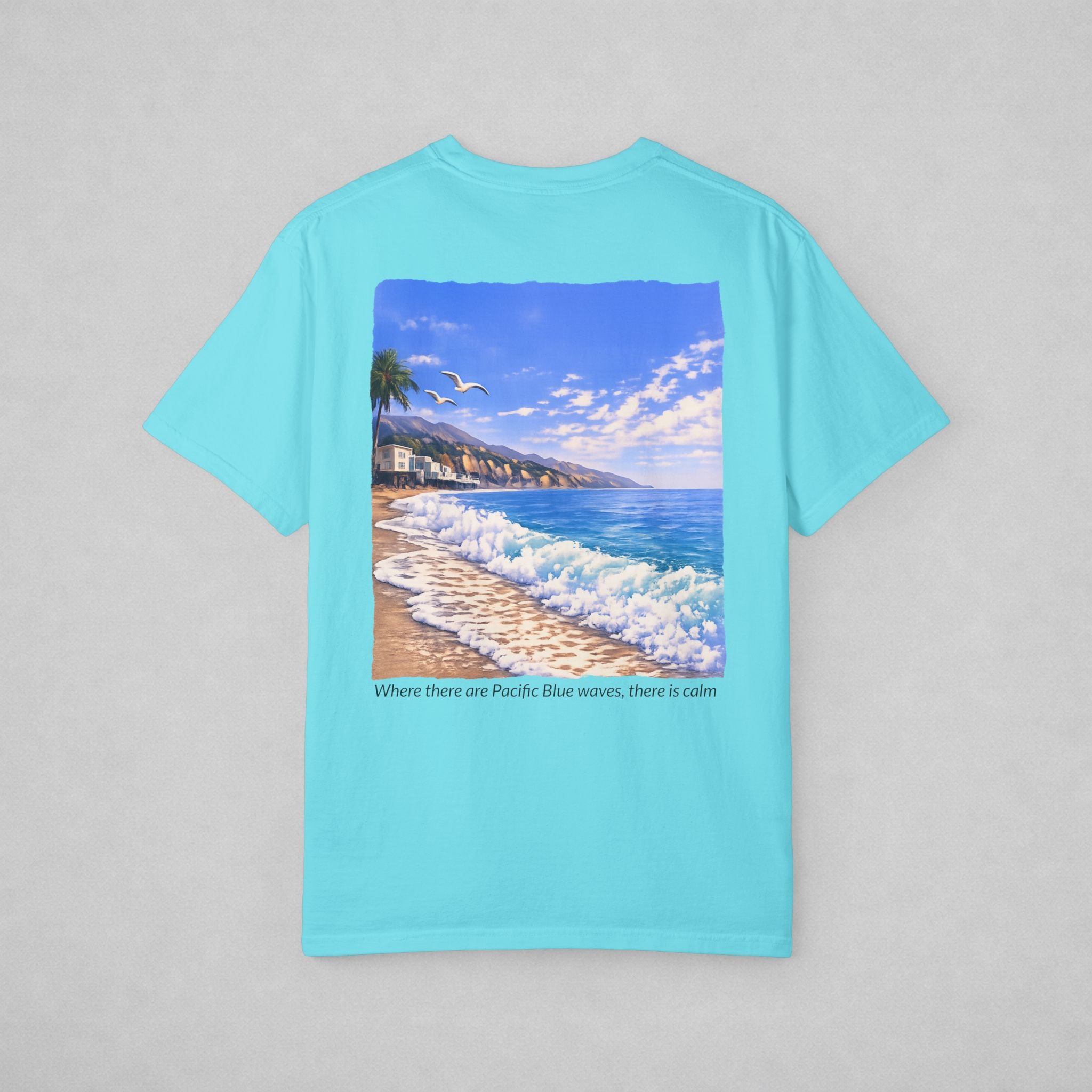 A light blue short-sleeve t-shirt with a large square graphic print on the back depicting a beach scene with blue waves, a sandy shore, and a coastal landscape under a blue sky. Below the graphic, the text reads 'Where there are Pacific Blue waves, there is calm'.
