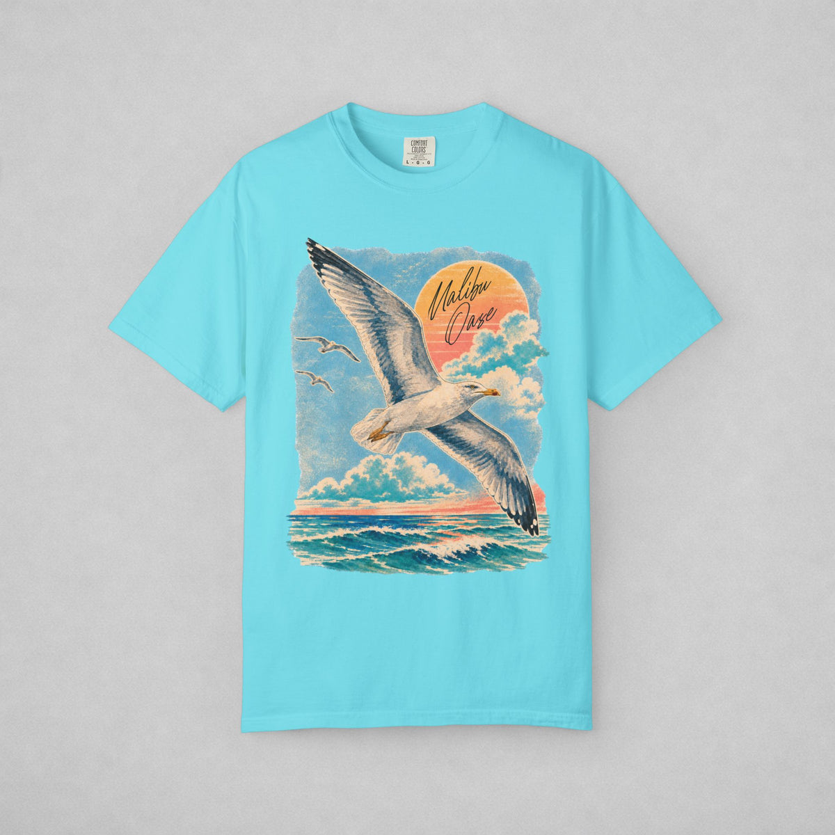 A light blue short-sleeve crewneck t-shirt featuring a large graphic print of a seagull flying over ocean waves under a sunset sky, with the words 'Malibu Oase' written in the sky.