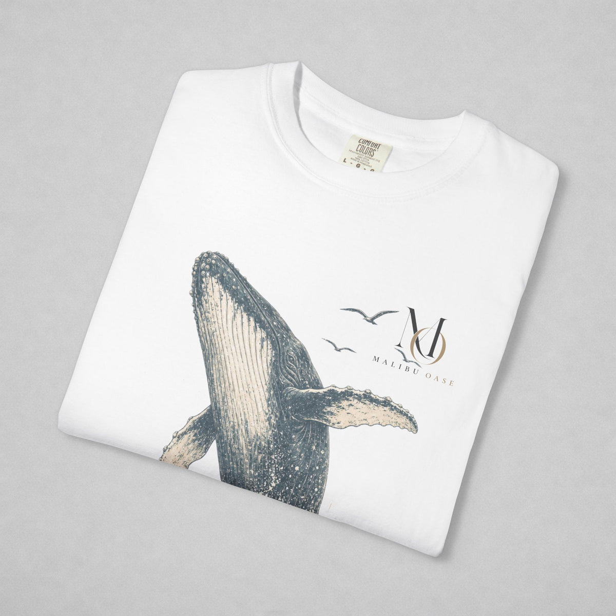 A folded white crewneck sweatshirt on a light gray background, featuring a graphic print of a whale, birds, and a stylized 'Malibu Oase' monogram.
