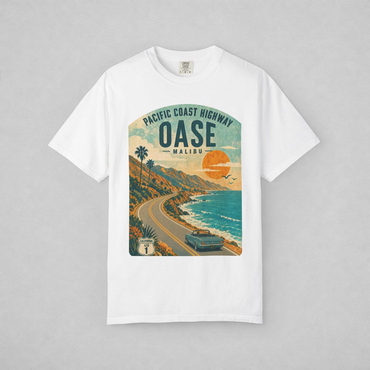 A white short-sleeve crewneck t-shirt with a vintage-style graphic print of the Pacific Coast Highway in Malibu, California. The graphic features a winding coastal road, palm trees, a car, and a large orange sun over blue ocean water, with 'OASE MALIBU' printed in bold text.