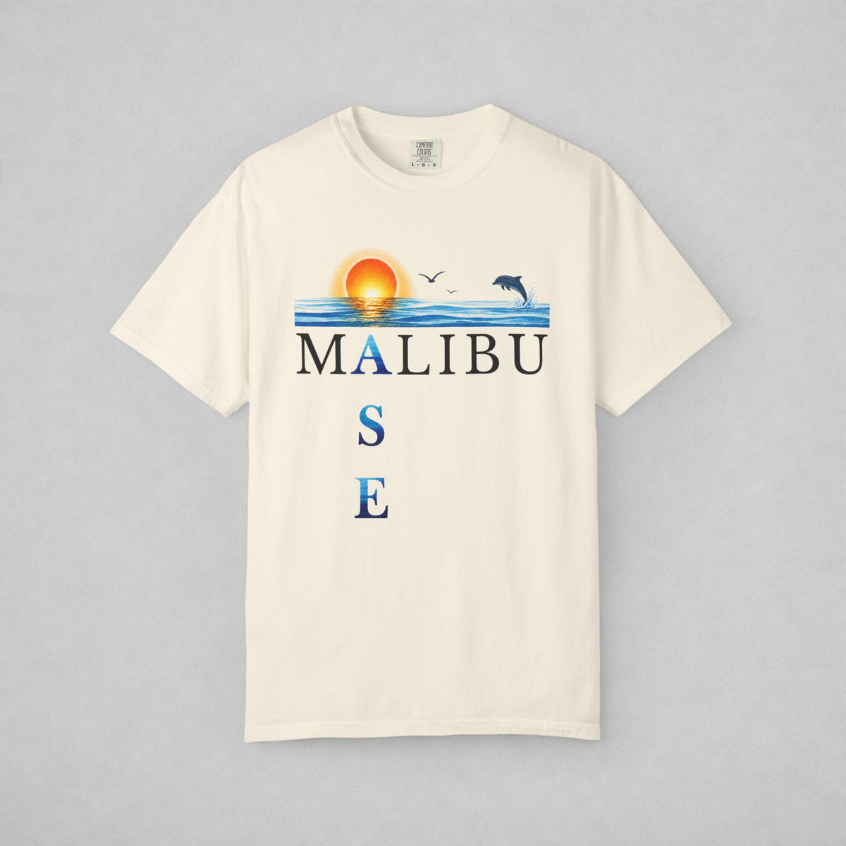 An off-white short-sleeve t-shirt with a graphic print on the front featuring a sunset over blue water, birds, a dolphin, and the text 'MALIBU OASE'. The shirt is displayed flat against a neutral gray background.