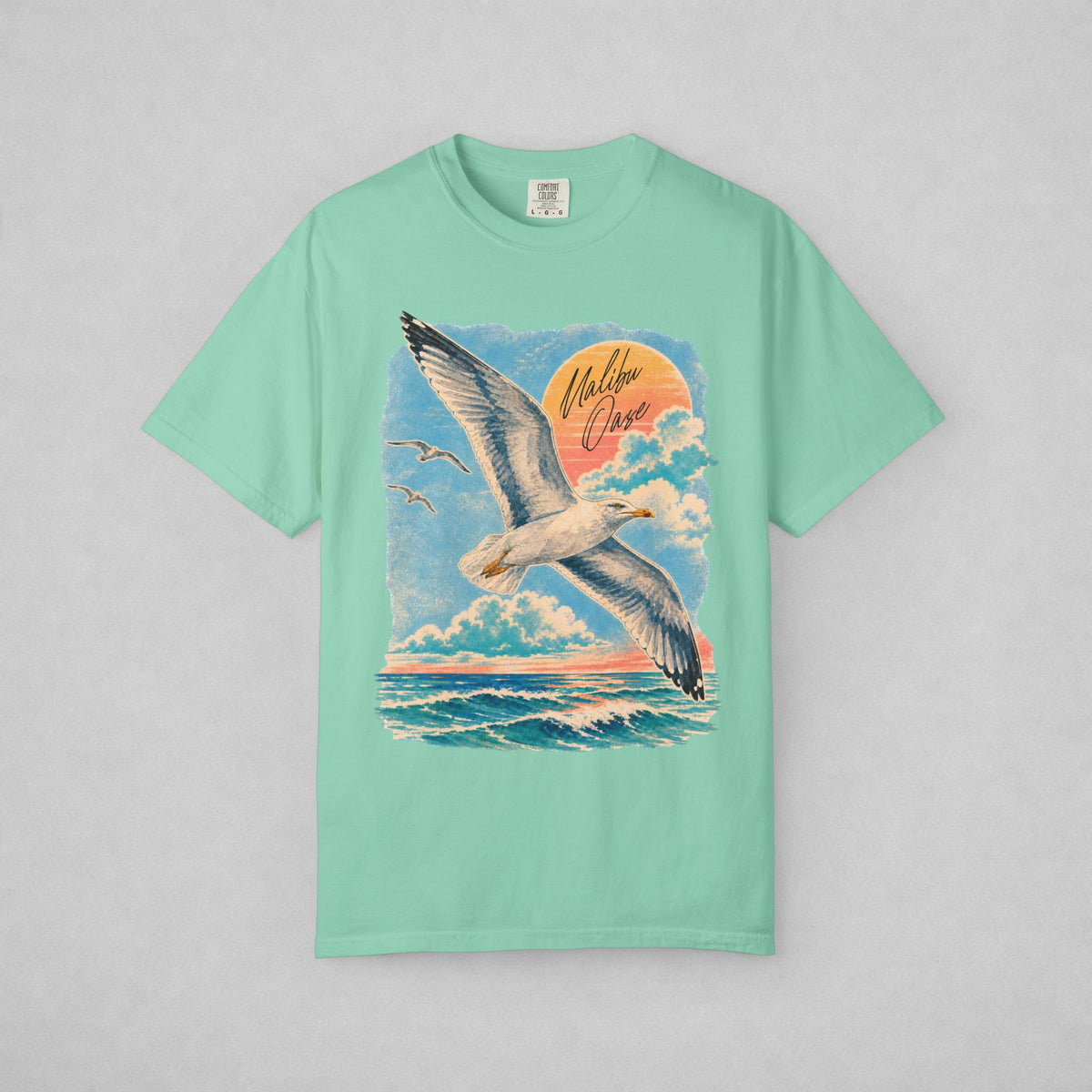 A mint green short-sleeve t-shirt with a large graphic print of a flying seagull over a blue ocean under a sunset sky, featuring the text 'Malibu Oase'. The shirt is displayed flat against a neutral gray background.