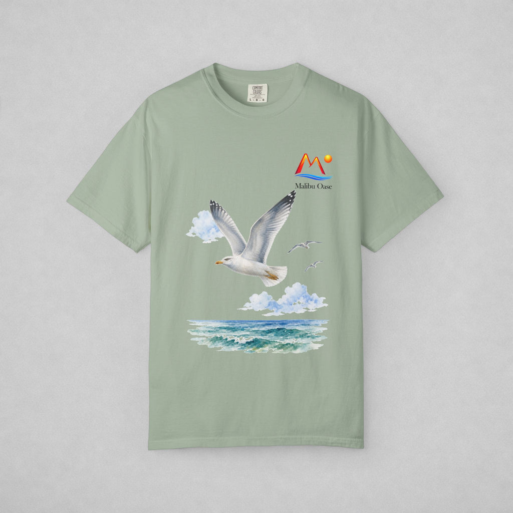 A sage green short-sleeve t-shirt with a graphic print of a flying seagull, clouds, ocean waves, and a 'Malibu Oase' logo on the chest, displayed flat against a neutral gray background.