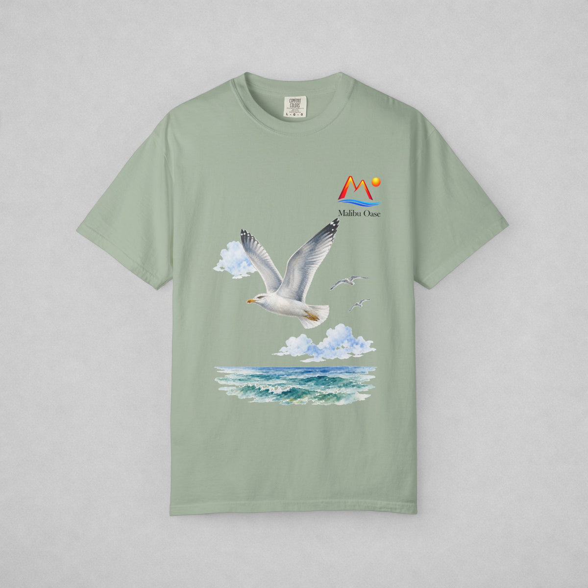 A sage green short-sleeve t-shirt with a graphic print of a flying seagull, clouds, ocean waves, and a 'Malibu Oase' logo on the chest, displayed flat against a neutral gray background.