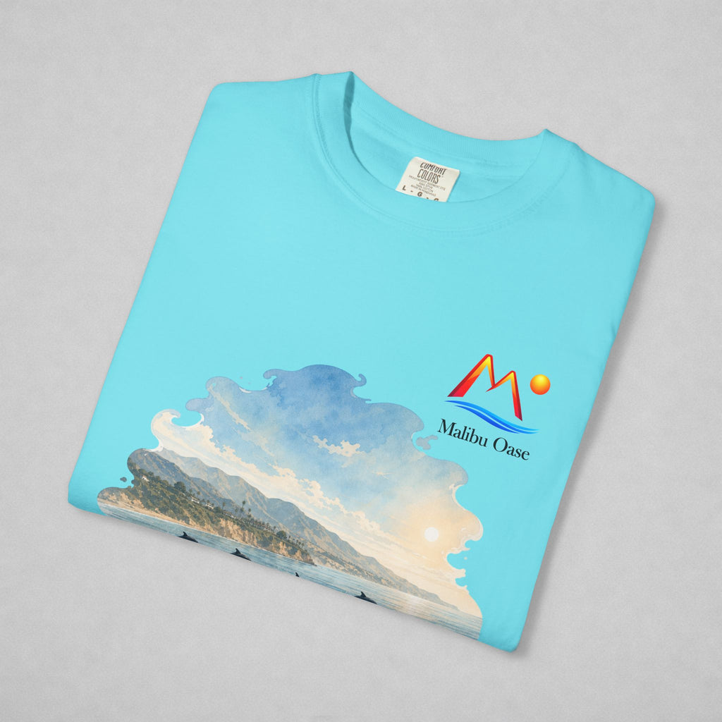 A folded Lagoon Blue t-shirt on a light gray background, featuring a colorful graphic print of a coastal landscape with mountains, waves, and a sun, alongside the 'Malibu Oase' logo.