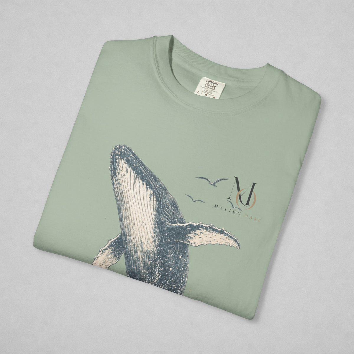 A folded sage green t-shirt with a whale graphic and 'Malibu Oase' logo printed on the front, displayed on a neutral gray background.