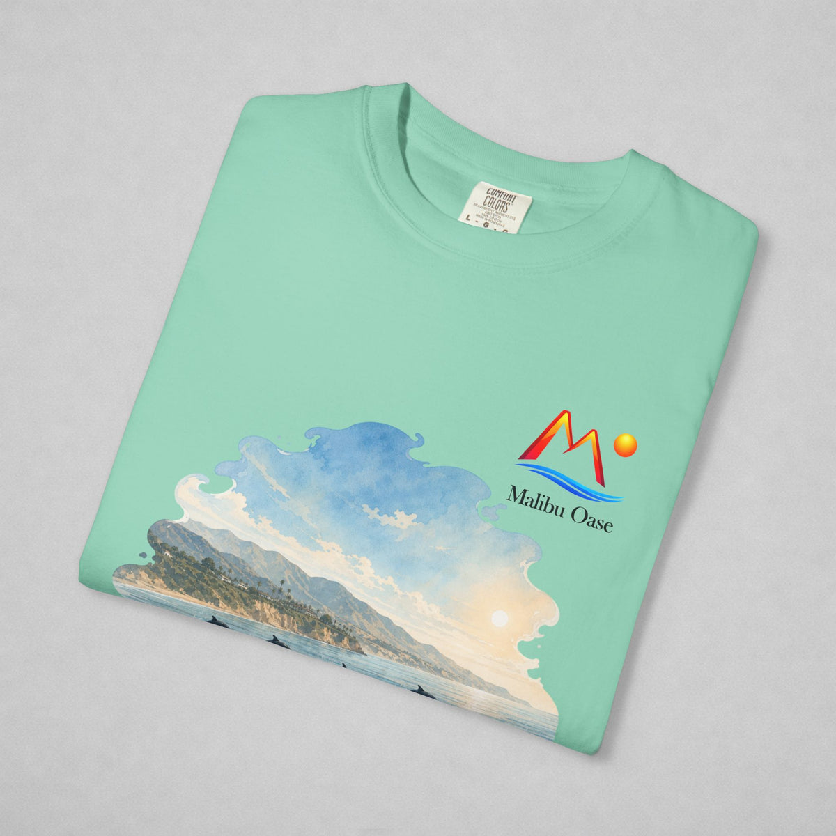 A folded Island Reef t-shirt on a light gray background, featuring a colorful graphic print of a coastal landscape with mountains, waves, and a sun, alongside the 'Malibu Oase' logo.