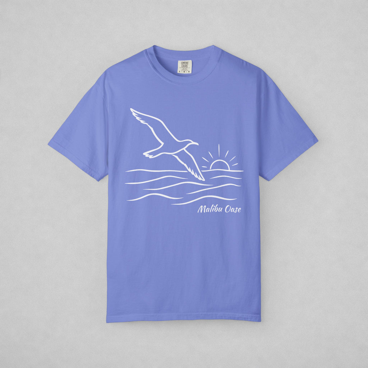 Flo Blue.  Short-sleeve t-shirt with a white line art graphic of a flying seagull, waves, a rising sun, and the text Malibu Oase' on the front, displayed flat against a neutral gray background.