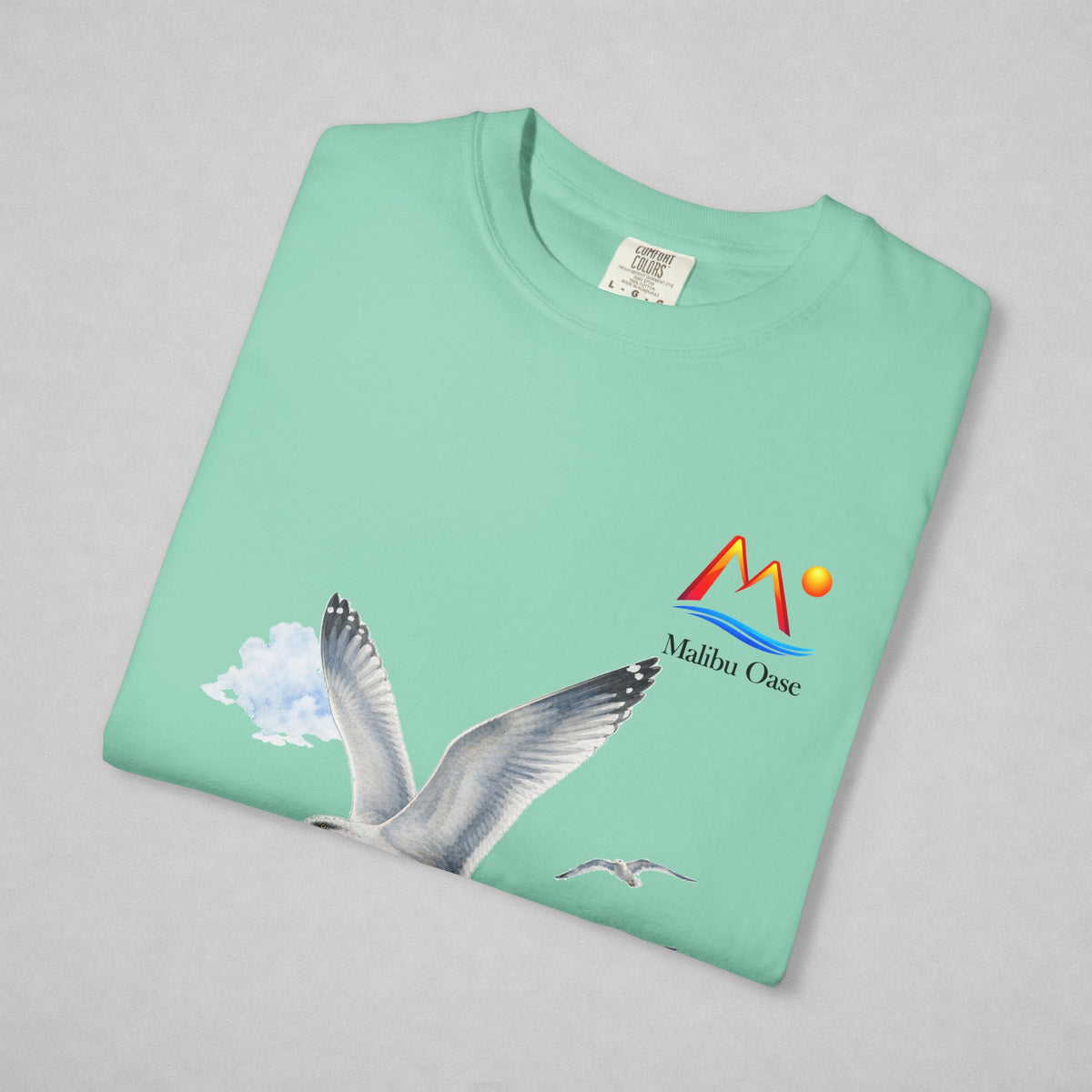 A folded mint green t-shirt on a light gray background, featuring a graphic of a flying seagull and a colorful 'Malibu Oase' logo on the chest.
