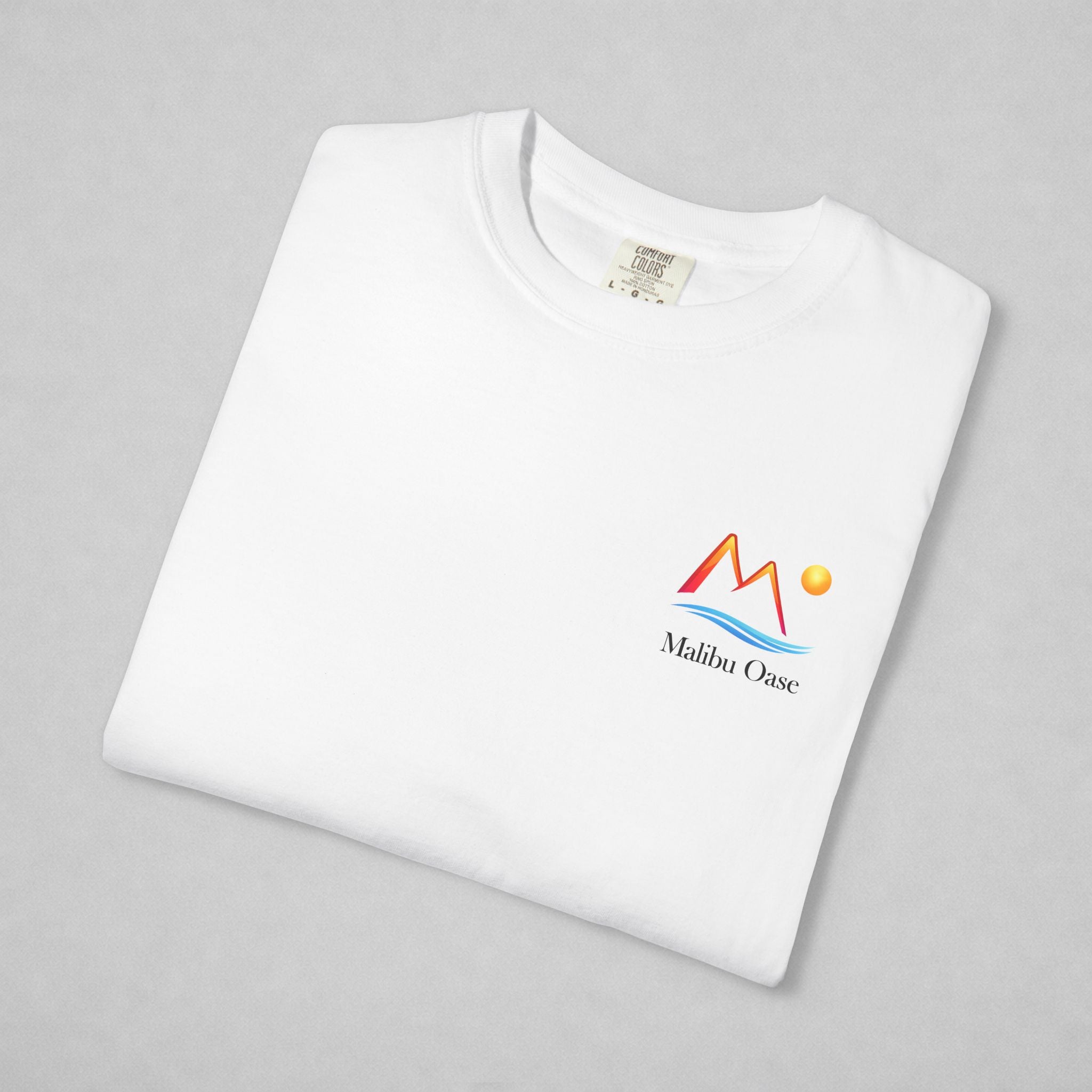 A folded white crewneck t-shirt on a light gray background, featuring a colorful 'Malibu Oase' logo printed on the left chest.