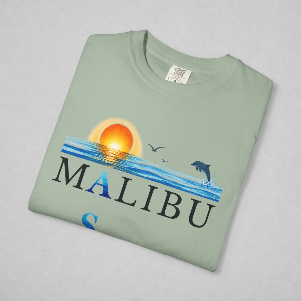 A folded sage green t-shirt with a 'MALIBU' ocean graphic print, featuring a sunset, waves, birds, and a dolphin, displayed on a neutral gray background.