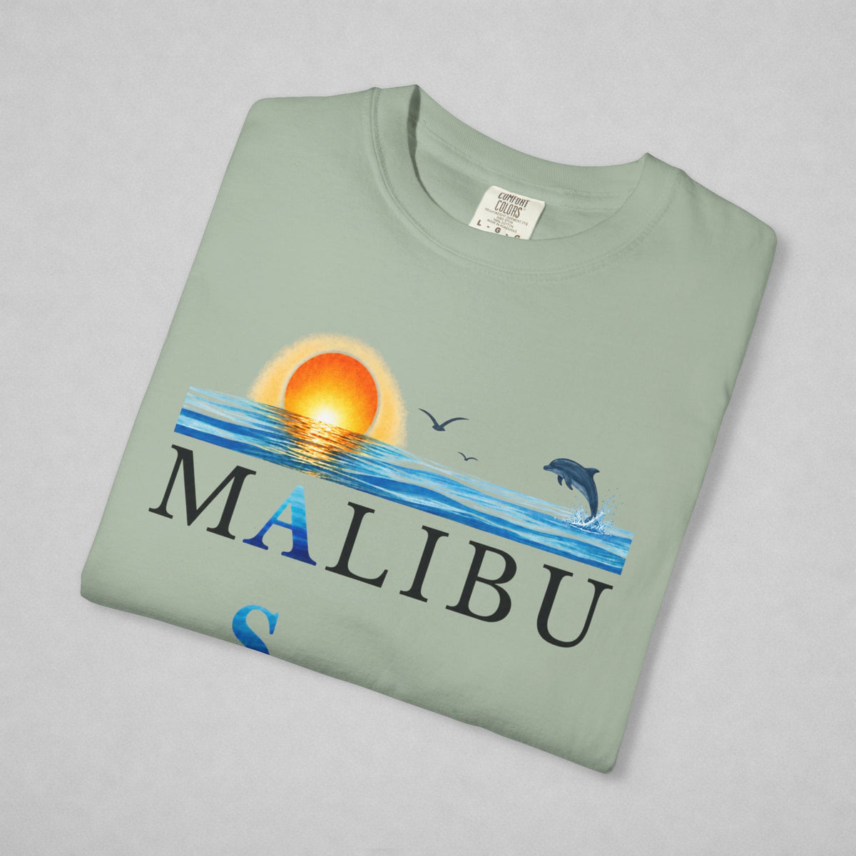 A folded sage green t-shirt with a 'MALIBU' ocean graphic print, featuring a sunset, waves, birds, and a dolphin, displayed on a neutral gray background.