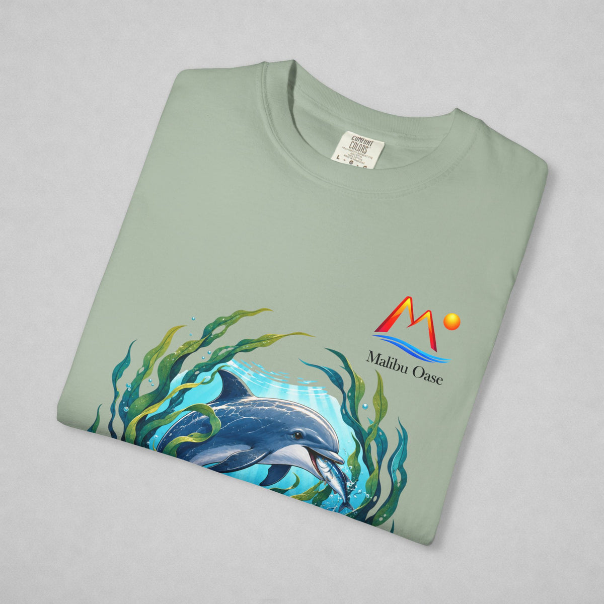A folded sage green t-shirt on a light gray background, featuring a colorful graphic of a dolphin swimming underwater with green seaweed and a 'Malibu Oase' logo printed on the chest.
