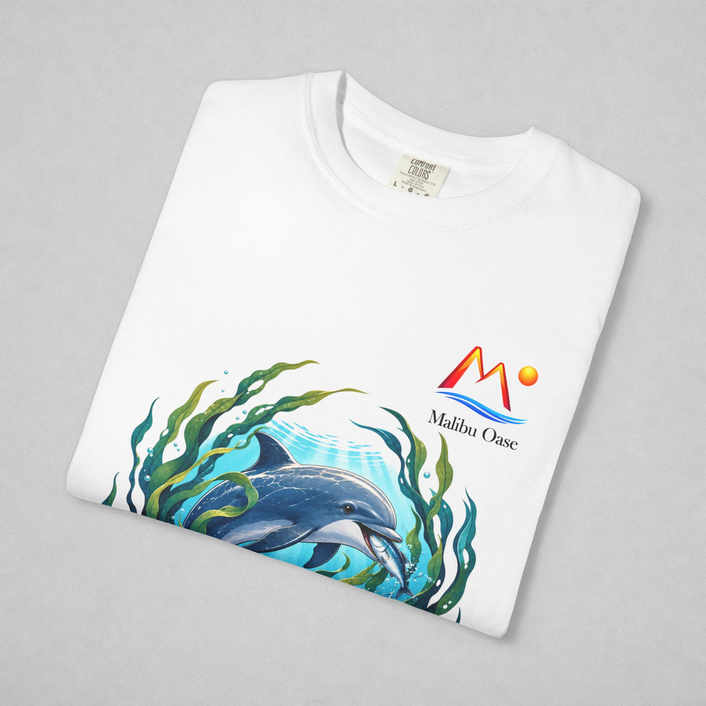A folded white t-shirt on a light gray background, featuring a colorful graphic of a dolphin swimming among green seaweed, with a red and yellow 'Malibu Oase' logo printed on the chest.