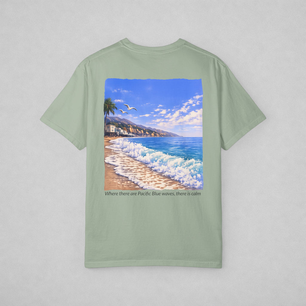 A sage green short-sleeve t-shirt with a large square graphic print of a beach scene featuring blue waves, a sandy shore, and a palm tree. Below the graphic, the text reads 'Where there are Pacific Blue waves, there is calm'.