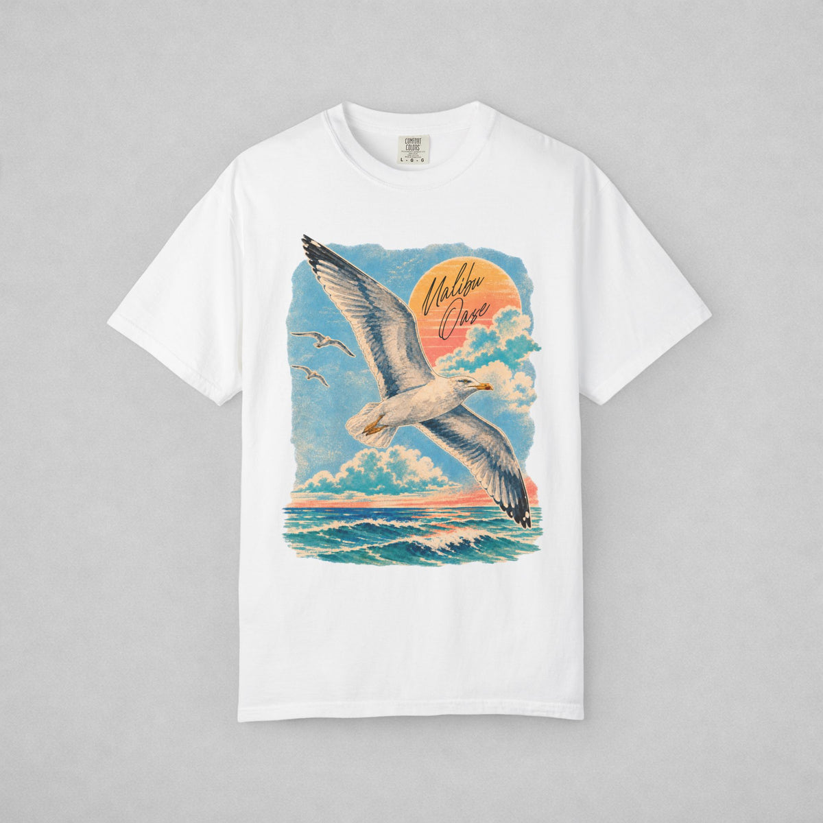 A white short-sleeve crewneck t-shirt featuring a large graphic print of a seagull flying over the ocean under a sunset sky, with the words 'Malibu Oase' written in the sky.