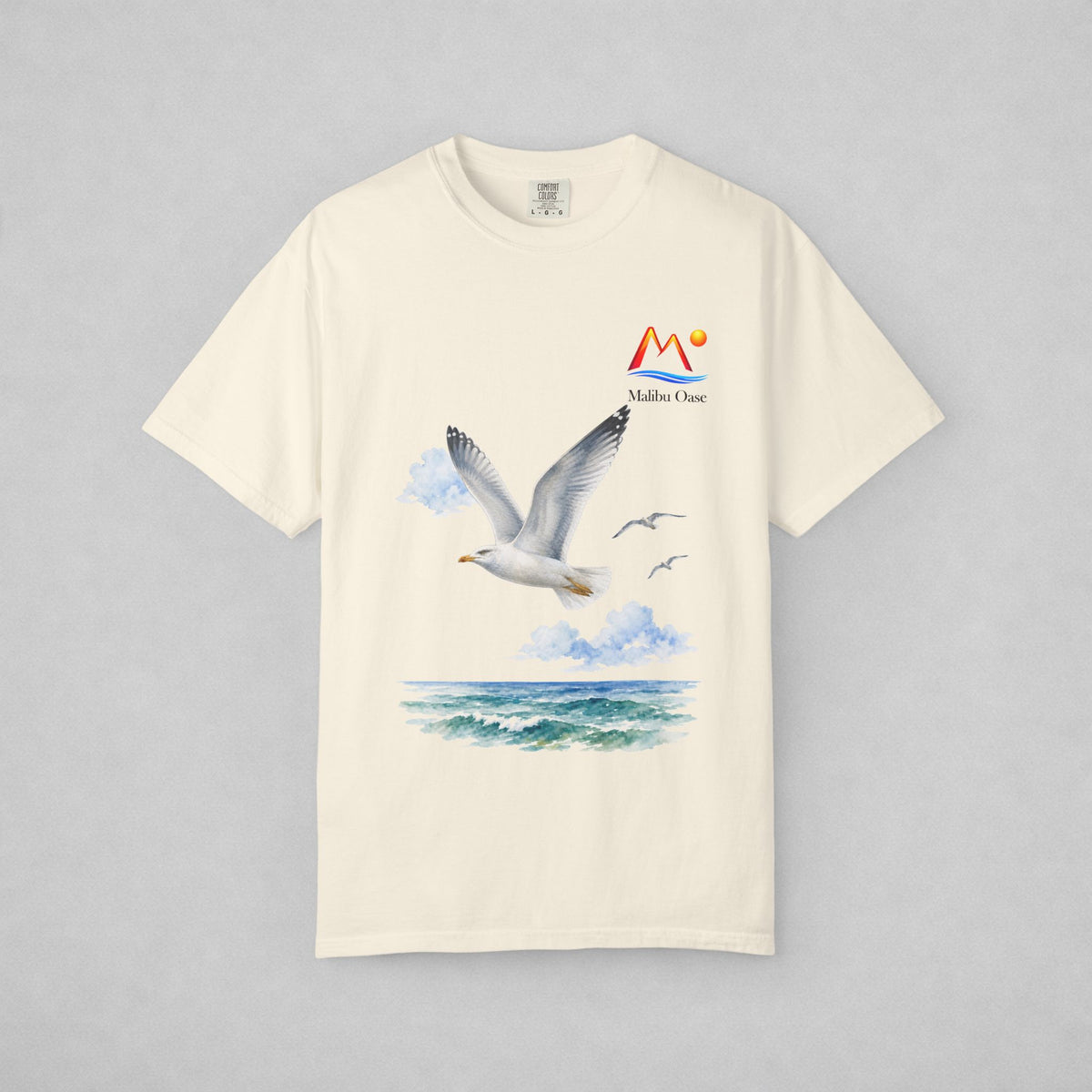 An off-white short-sleeve t-shirt with a graphic print of a flying seagull over blue ocean waves, featuring a 'Malibu Oase' logo on the right chest, displayed on a neutral gray background.
