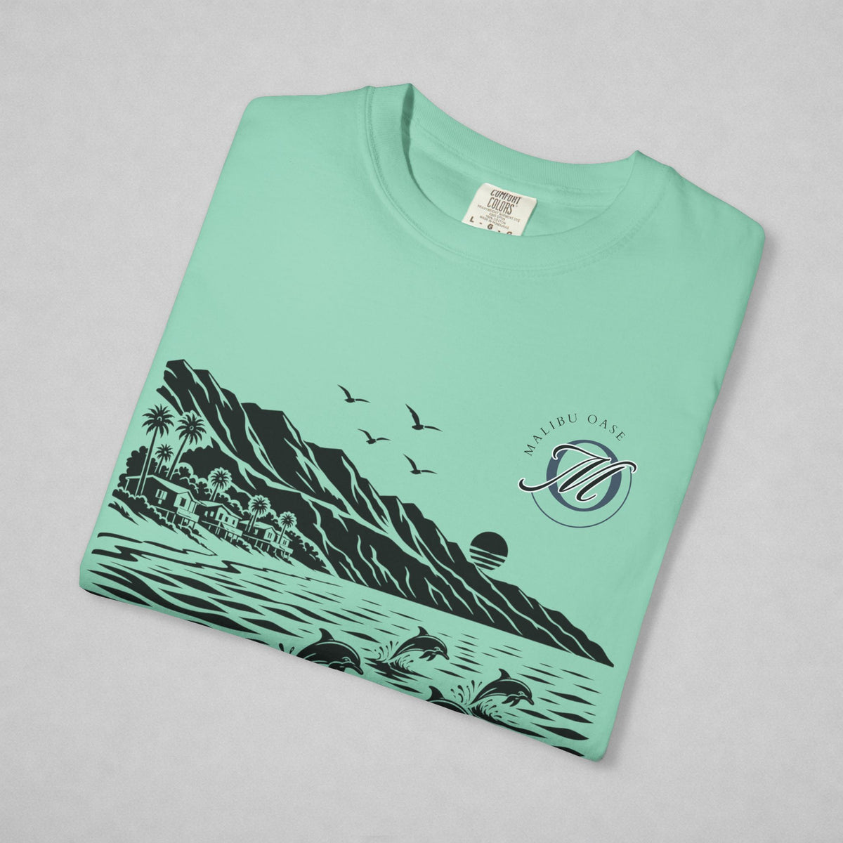 A mint green folded t-shirt with a black graphic print of a coastal landscape, including mountains, palm trees, birds, dolphins, and a sunset. The graphic also features the 'Malibu Oase' monogram.