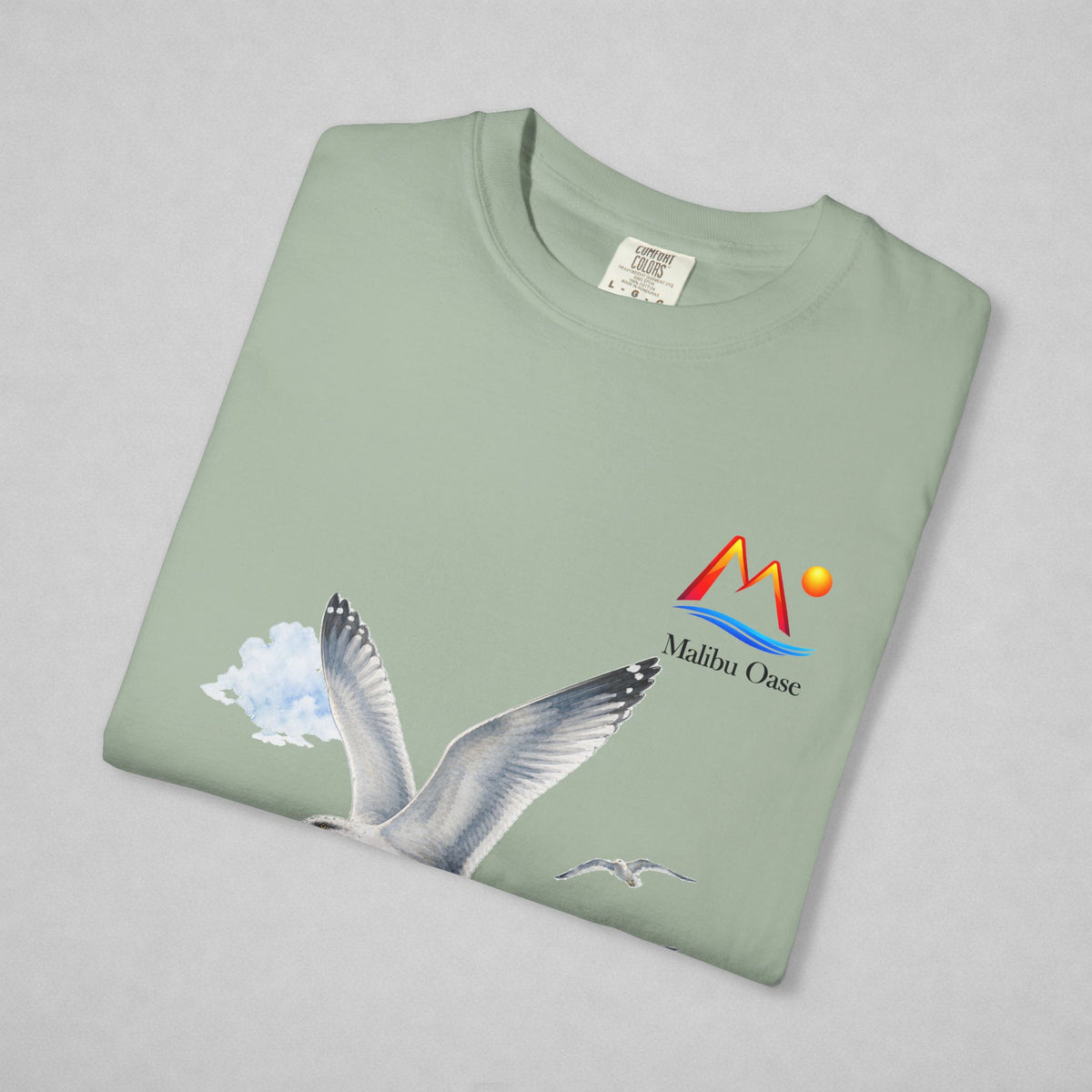 A folded sage green t-shirt on a light gray background, featuring a graphic of a flying seagull and a colorful 'Malibu Oase' logo on the chest.