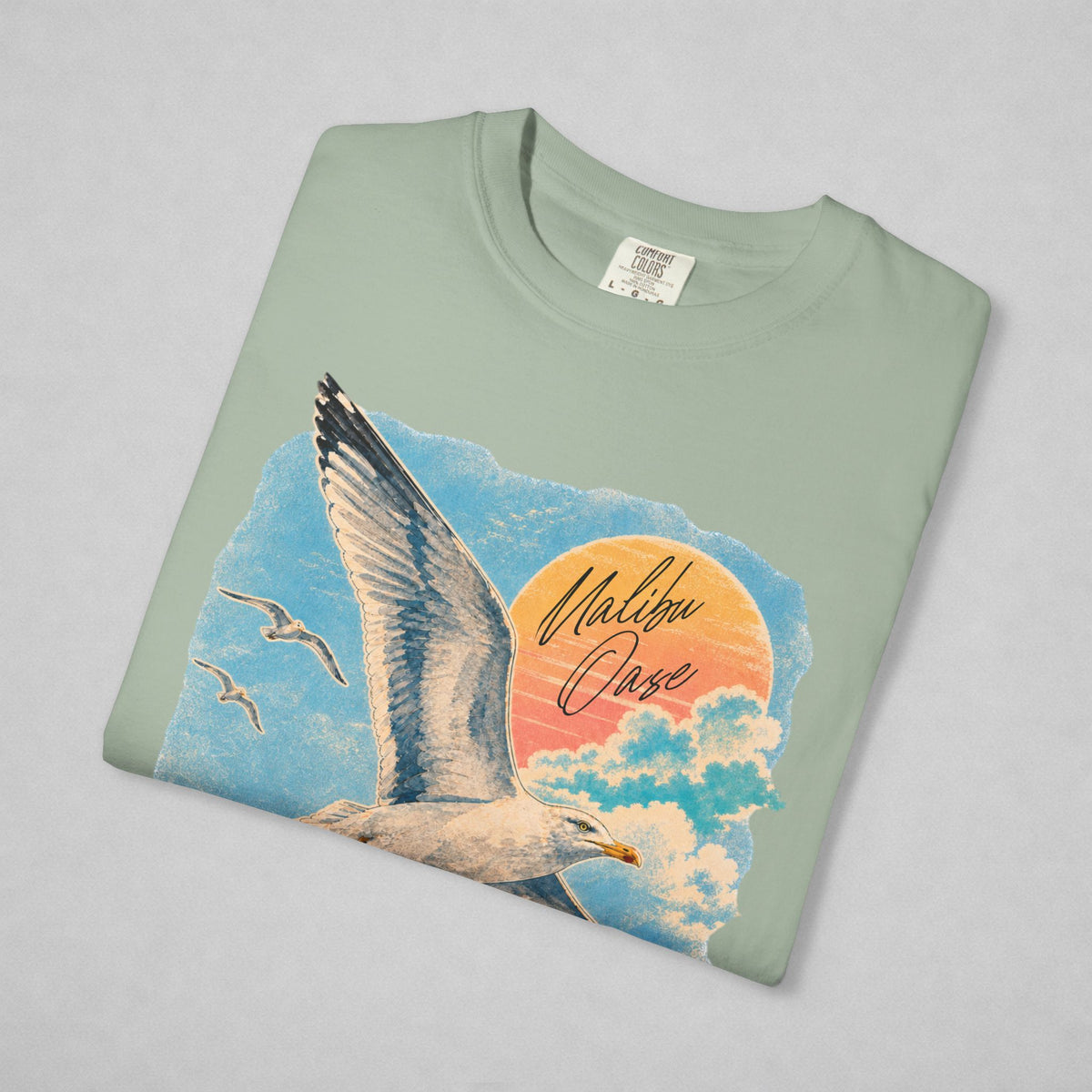 A folded sage green t-shirt with a colorful graphic print of a seagull, clouds, and a sunset with the text 'Malibu Oase', displayed on a neutral gray background.
