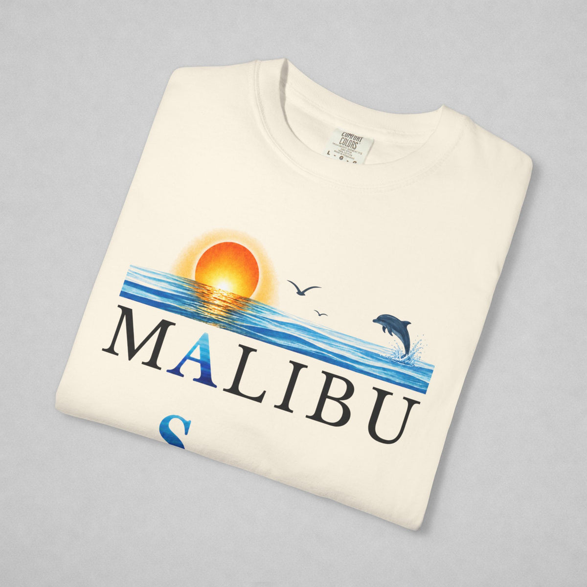 An off-white folded t-shirt featuring a colorful graphic print of a sunset over ocean waves with a jumping dolphin, seagulls, and the word 'MALIBU' printed across the front, displayed on a neutral gray background.