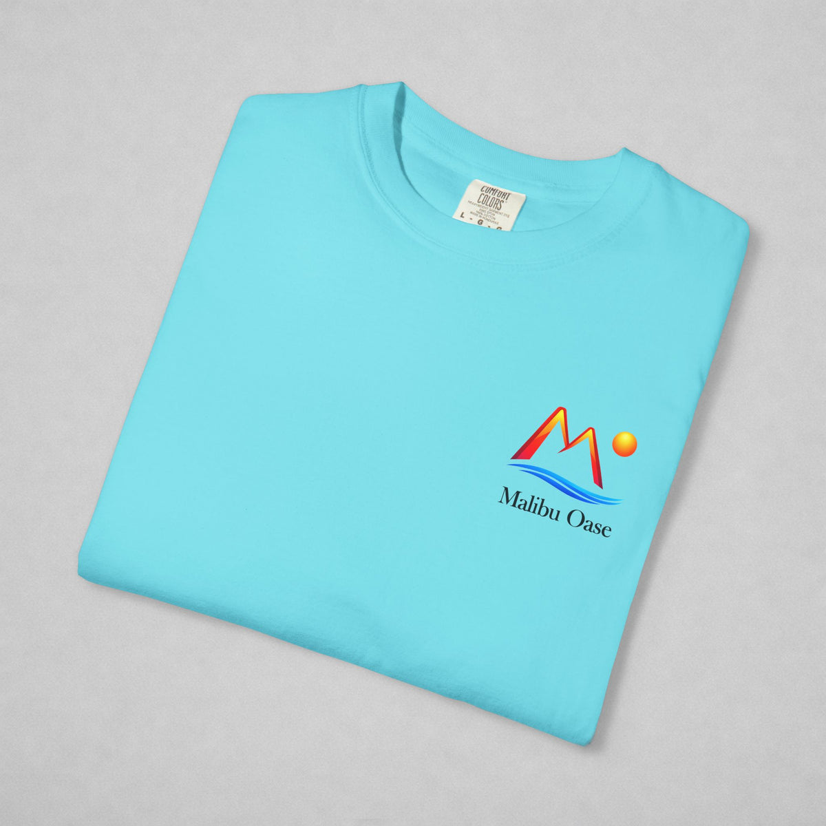 A folded light blue t-shirt on a neutral gray background, featuring a colorful 'Malibu Oase' graphic on the left chest with red, orange, yellow, and blue elements.