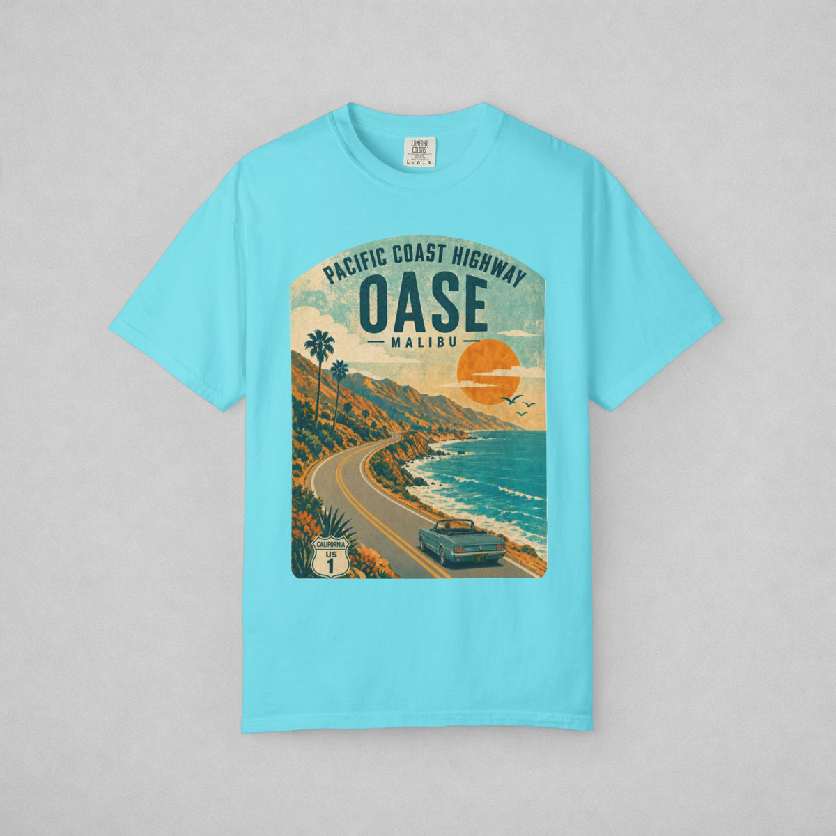 A light blue short-sleeve t-shirt with a vintage-style graphic print of the Pacific Coast Highway in Malibu. The graphic features a winding coastal road, palm trees, a car, and a large orange sun over the ocean, set against a neutral gray background.