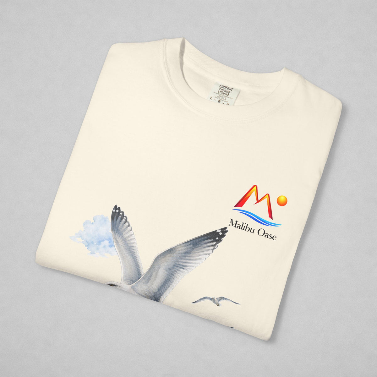 A folded cream-colored t-shirt on a light gray background, featuring a graphic of a flying seagull in blue and gray, and a colorful 'Malibu Oase' logo on the left chest.