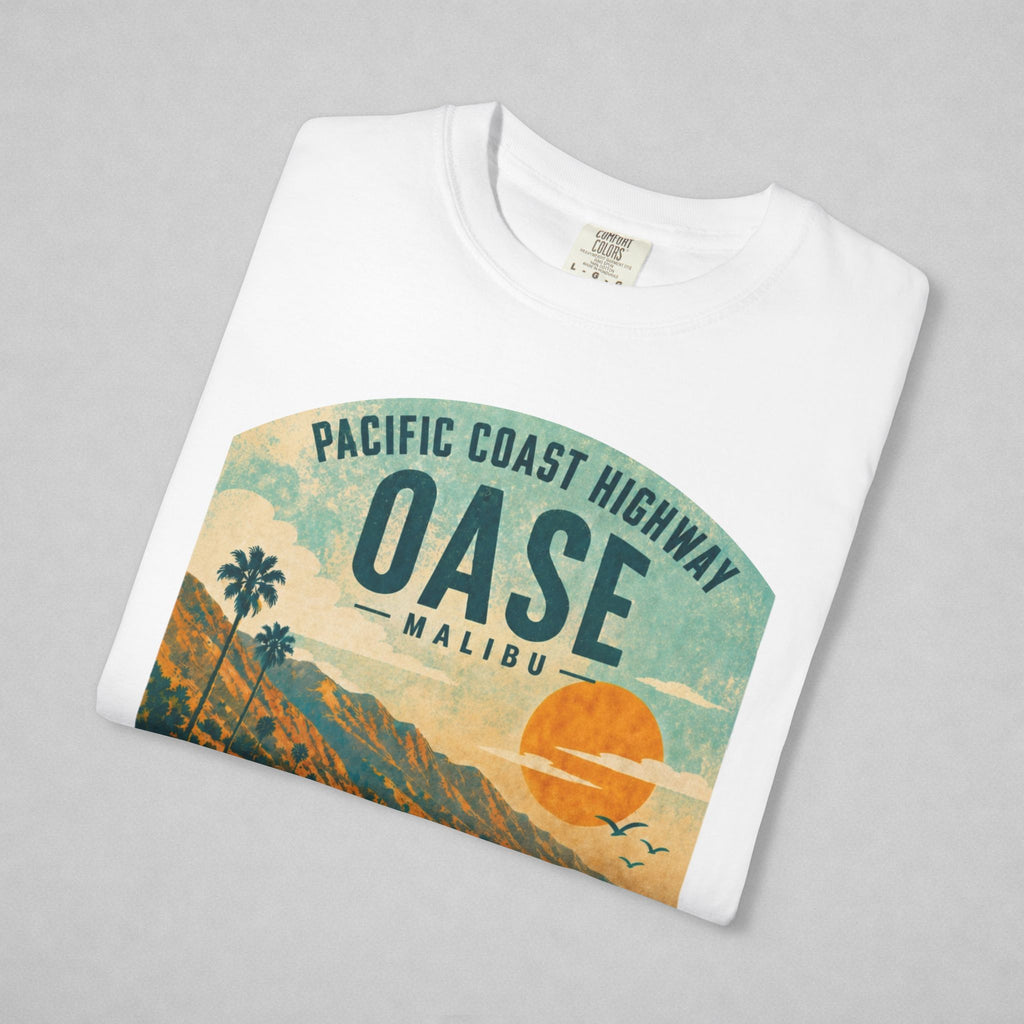 A folded white crewneck t-shirt with a retro graphic print of the 'Pacific Coast Highway Oase Malibu' scene, displayed on a neutral gray background.