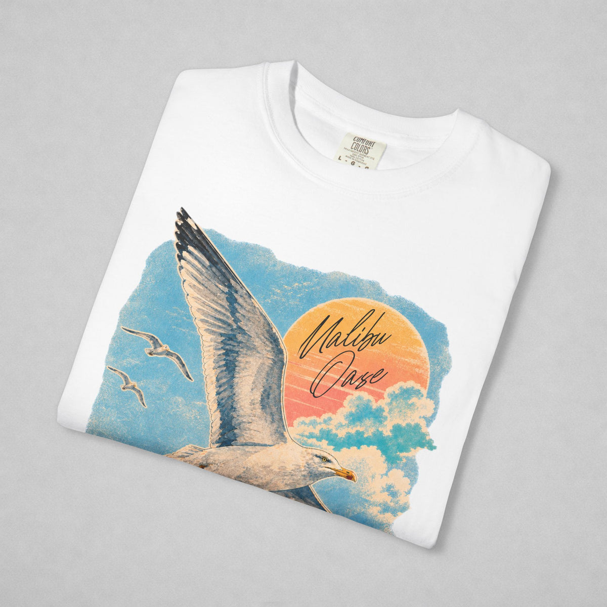A folded white crewneck t-shirt with a colorful graphic print of a seagull flying over clouds and a sunset, featuring the text 'Malibu Oase' in script, displayed on a neutral gray background.